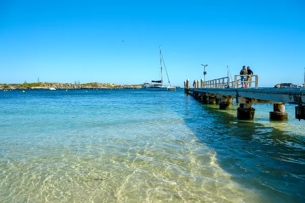 A sunny beach with a wooden pier extending into the blue ocean. A sailboat is anchored near the end of the pier, and two people are standing on it. 