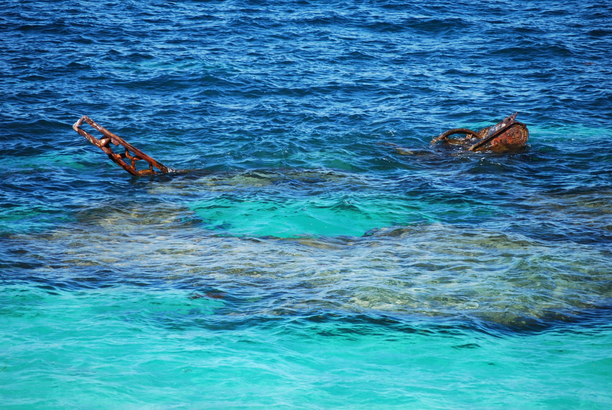 The remains of a shipwreck partially submerged in clear blue ocean water, with rusted metal visible above the surface.