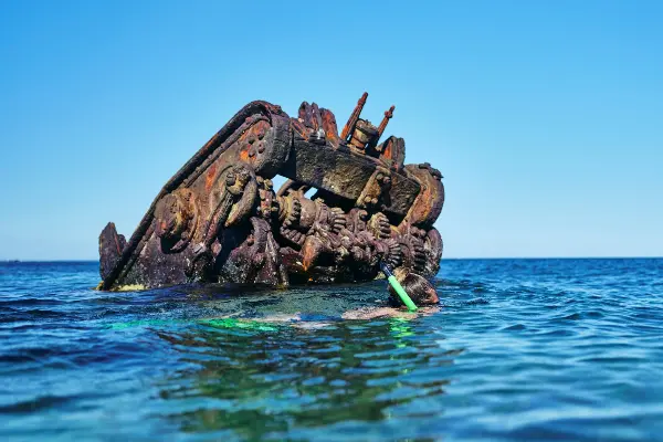 A person snorkeling near a partially submerged, rusted shipwreck in clear blue water under a bright blue sky.