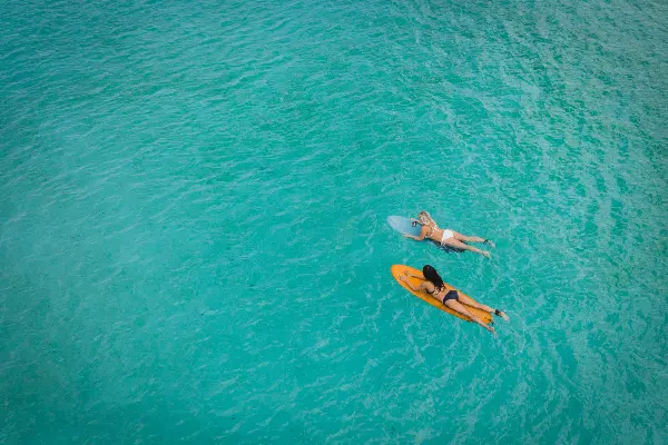 Two people lying on surfboards in clear turquoise water, viewed from above. One person is on an orange surfboard, and the other is on a blue surfboard.