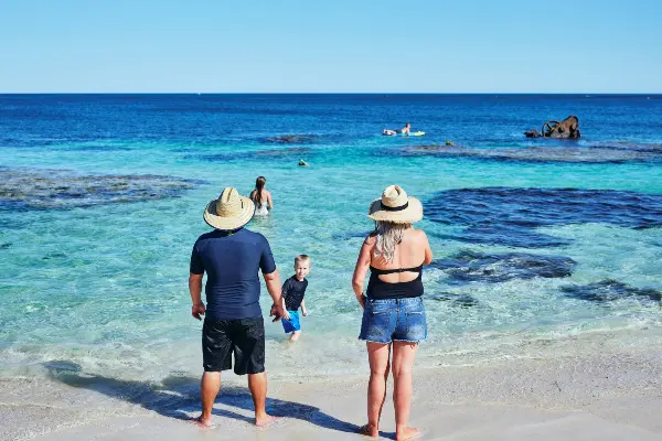 Two people in straw hats stand on a sandy beach, facing the ocean. A child in blue swimwear walks towards the water. The ocean is clear with some rocks visible, and two more people are swimming near the rocks under a clear blue sky.