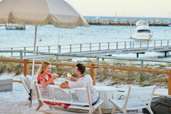 Couple looking at each other smiling sitting under an umbrella in the evening having a drink with views of Thomson Bay