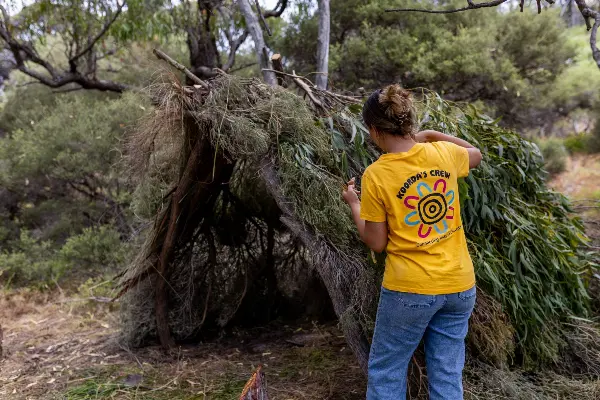 A woman with a yellow shirt on building a shelter out of branches
