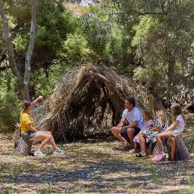 Family sitting looking at a shelter built by branches surrounded by trees