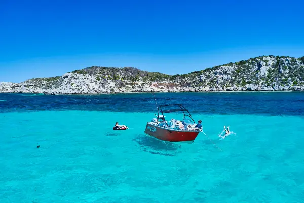 A red boat is anchored in clear, turquoise water with a few people swimming around it. The background features a rocky shoreline with sparse vegetation under a bright blue sky.