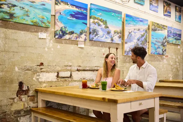 Couple eating at Geordie's Cafe & Art Gallery with art prints on wall behind them of Rottnest Island beaches
