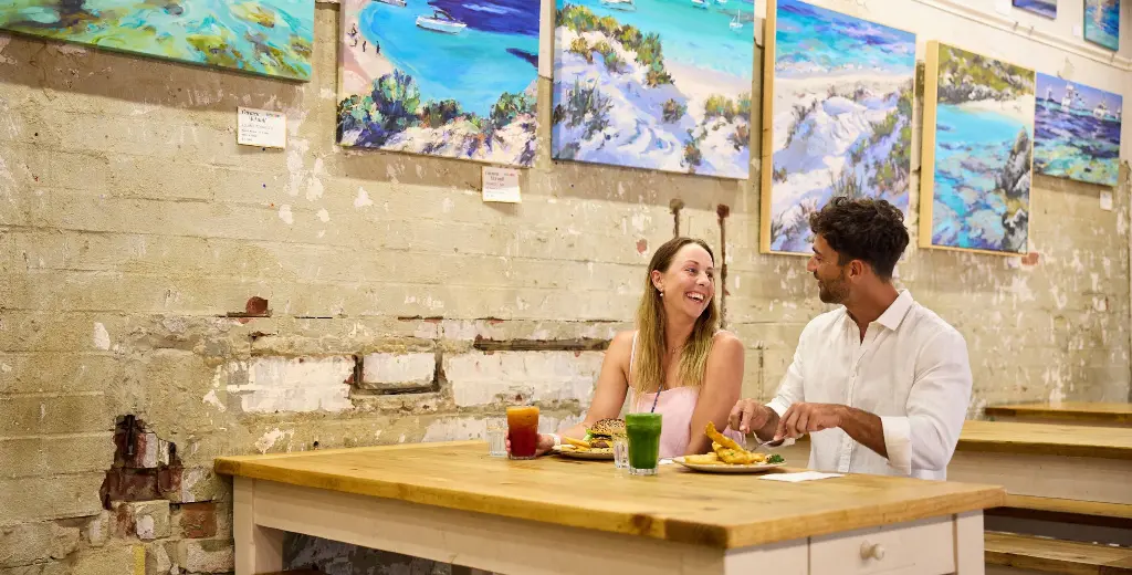 Couple eating at Geordie's Cafe & Art Gallery with art prints on wall behind them of Rottnest Island beaches