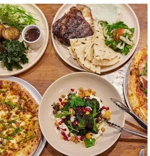 Two Pizzas, a steak and other dishes on a table.