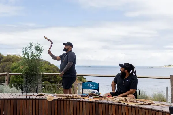 Two men sitting in front of the ocean, one holding a boomerang and the other sitting looking at him smiling